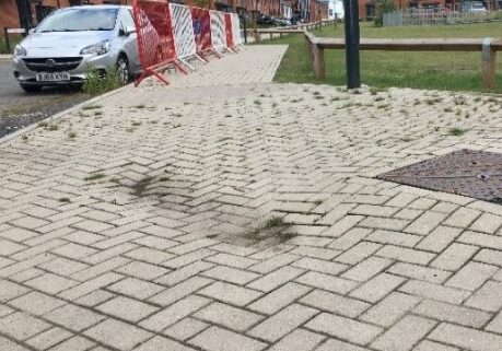 Pale buff block paving which has subsided near a utility cover creating pothole areas where water and debris puddle. There is also significant weed growth visible on the block paving in the background