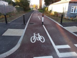 Photo of a pavement and cycle path. The pavement is kerbed above the level of the cycle path. There is a buff coloured tactile crossing for pedestrians to cross the cycle path. Four bollards spaced with 1.5m gaps prevent vehicle access. The cycle path is red and the pavement grey/black asphalt.