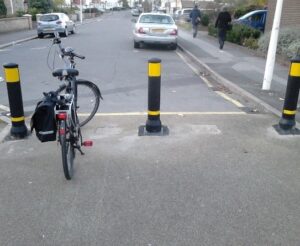 Photo shows a cycle access across a pavement onto a residential street. All surfaces are grey asphalt. There are three black bollards with bright yellow reflective bands spaced with 1.5m gaps between them positioned near the edge of the drop kerb. There is a bicycle standing in one of the gaps between the bollards. It has no rider- it is probably the photographer's bike.