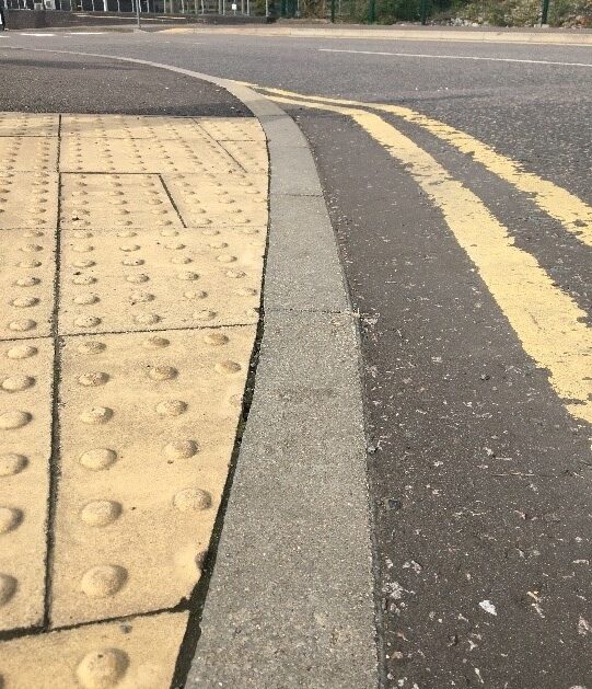 Photo of part of an uncontrolled road crossing has buff tactile paving to the left, a fully flush pale grey concrete kerb in the middle then dark asphalt with double yellow lines to the right.