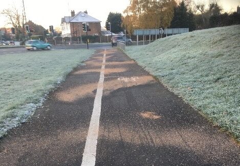 A traffic-free asphalt path with cycle and pedestrian sides demarcated with a white thermoplastic paint line. There is frosty grass to either side of the path, a road visible in the distance and pale brown grit scattered across the path surface.