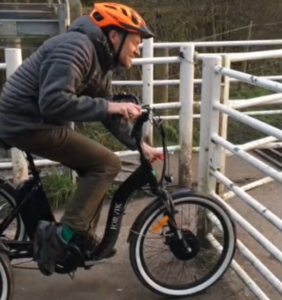 Photo shows a white man wearing an orange cycle helmet and black coat riding an upright Jorvik tandem. His front wheel is against a white metal gate and he is unsuccessfully reaching towards the gate lock with a RADAR key in his left hand.