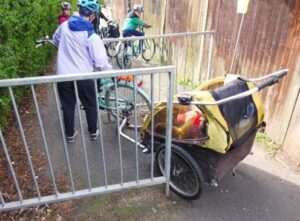 Photo shows a chicane made from two metal railings in an alleyway. There is a hedge to the left side and a solid wooden fence to the right. A bicycle with a child trailer attached is stuck in the chicane. The rider has dismounted and is attempting to free it.
