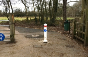 Photo shows the same location as photo above, photographed from the other side of the gates. The farm style gate is still visible to the left side of the photo. Where the kissing gate was, there is now a single white bollard with a red reflective band and blue cycle/pedestrian shared use circle on it.