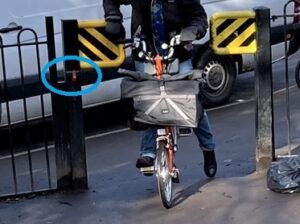 Photo shows a metal P barrier in a narrow gap in metal fencing. A cyclist on a Brompton is struggling to get through the gap which is on a steep hill. A white van behind the cyclist is also blocking the drop kerb access to the P barrier. A blue circle on the photo highlights the small red RADAR padlock which riders of many cycles and mobility aids will have to unlock in order to pass through the barrier.
