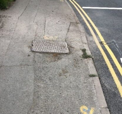 Photo showing asphalt footway to left and carriageway surface to right. The footway is badly worn, uneven and potholed with a utility cover creating a trip hazard. The carriageway has been newly resurfaced with smooth continuous asphalt.