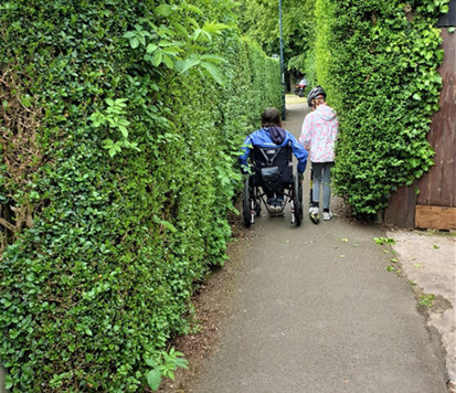 A wheelchair user and a child on a push scooter are just managing to squeeze down a narrow path between large privet hedges. The hedges are neatly cut but overhang the path almost to the point that the path is unusable.
