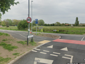 Google Streetview screenshot shows road junction with separate cycle and pedestrian paths crossing the road. The road is raised to the level of the pavement and give way signs apply to vehicles on carriageway. The pedestrian crossing has buff tactile paving. The cycle path is coloured red where the carriageway crosses it. There are no barriers on the pavement, cycle path or carriageway.