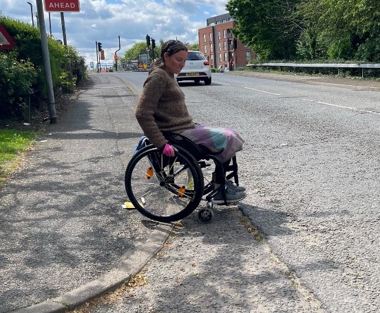 A manual active wheelchair user is cautiously edging down a steep, uneven drop kerb onto a broken tarmac road surface. The angle of the drop kerb means the wheelchair user is moving onto a busy road. The drop kerb has no tactile paving.