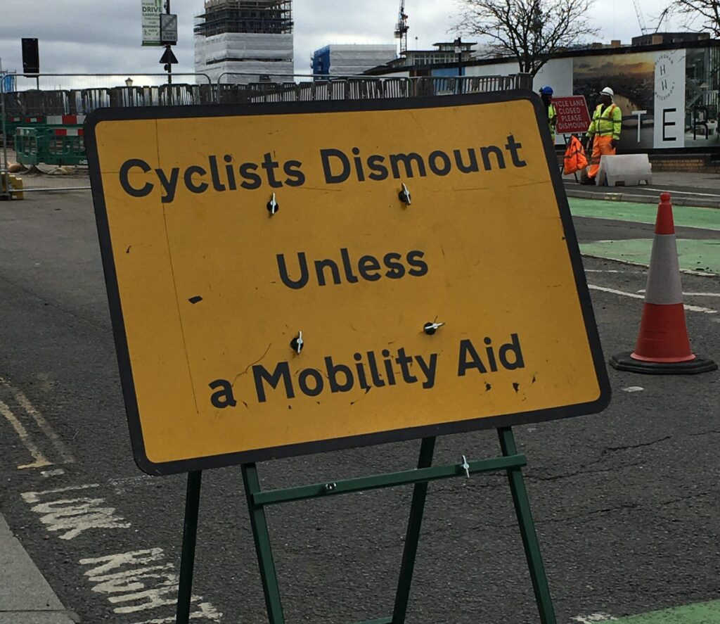 Yellow sign with black text that reads "Cyclists Dismount Unless Mobility Aid"