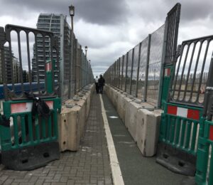 Barriers through bridge works: The path narrows to 1.2m between large concrete blocks with high fencing on either side. There is a white ridge of paint down the middle of the path.