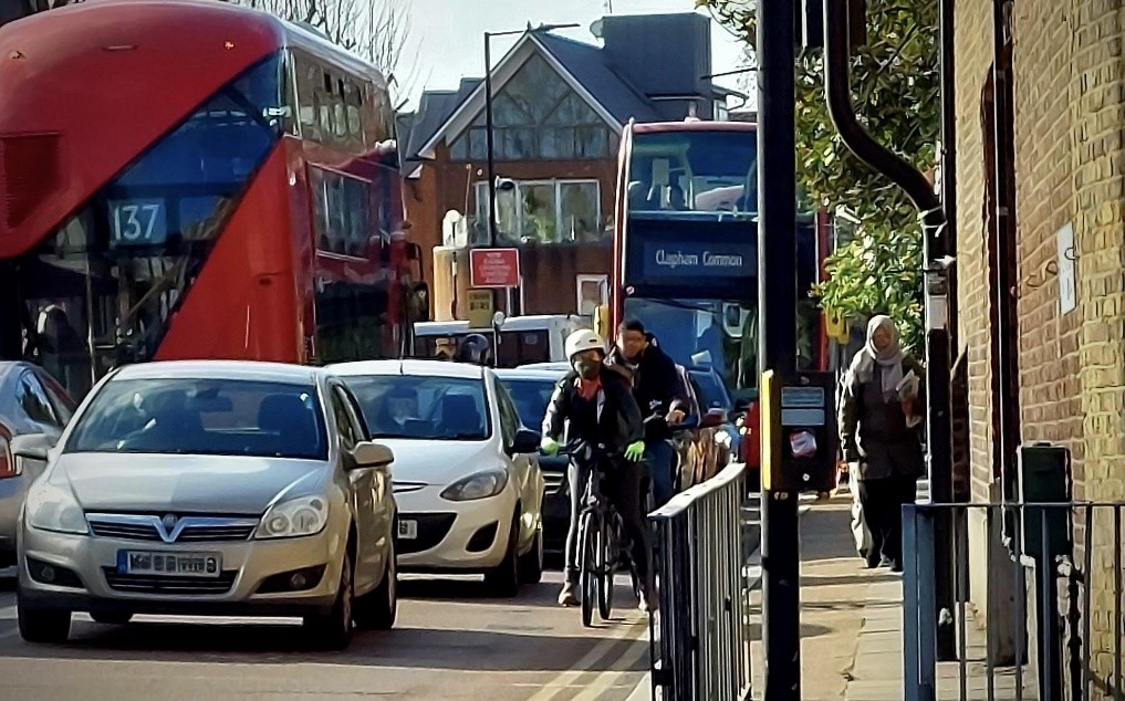 Photograph taken from a pavement showing oncoming traffic heading towards a junction. There are three cars with two cyclists on the inside of them and a double decker bus at the rear. A woman in Islamic dress is walking on the pavement.