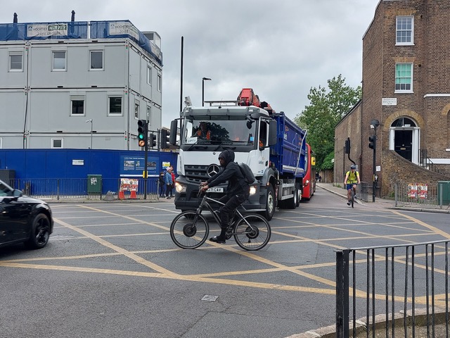 Photograph showing the centre of a large cross-roads junction. A large lorry is approaching in the oncoming direction while a cyclist cycles across the junction from right to left.