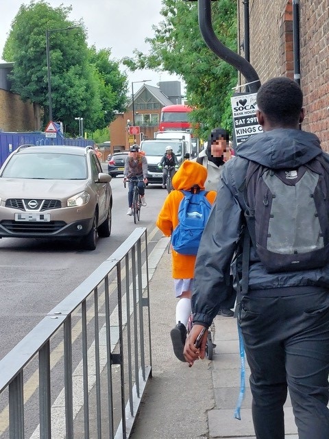 another photograph taken from the pavement facing the oncoming traffic at the junction. There are three people on the pavement and two cars and two cyclists approaching the camera.