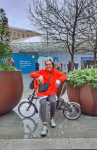 A smiling Black woman wearing a red jumper sitting sideways on a Brompton bike in between two big planters