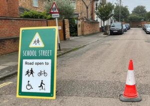 in foreground, on the road surface, a green sign saying “School street, Road open to” with graphics of children walking, a bicycle, a wheelchair user disability symbol and a person on a scooter. Also a traffic cone. A little behind them, a standard “school” road warning sign on a kerbside post. In the background rows of parked cars on both sides of the street, bay frontages of Victorian houses and a wall across the course of the road, showing where it changes direction. 