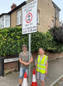 Cllr Lucy Bywater and Cllr Paul Edmonds, both smiling, with formal traffic sign saying “Pedestrian and cycle zone, Mon-Fri 8.15-9.00am 3.00-3.45pm”, and street name sign lower down “York Street”. In the background a house with rendered walls, and a large hedge. 