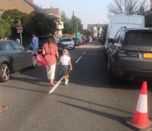 In foreground a traffic cone, a parent and child walking away from the camera down the middle of the road between parked cars. In middle distance a standard “school” road warning sign on a kerbside post with two adults close to it. In the distance further adults and children walking in the road, more parked cars and a Victorian house across the course of the road, showing where it changes direction.