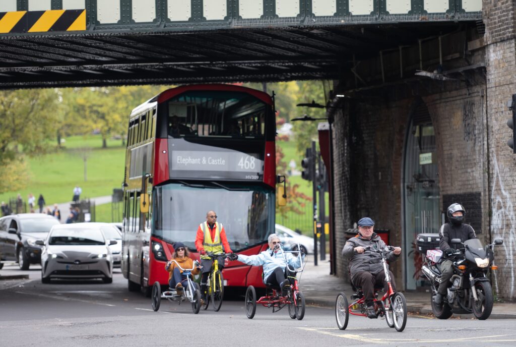 Four cyclists, three on recumbent trikes and one on a bicycle, entering a junction under a rail bridge with a motorcyclist to their left and a double-decker bus and traffic behind.