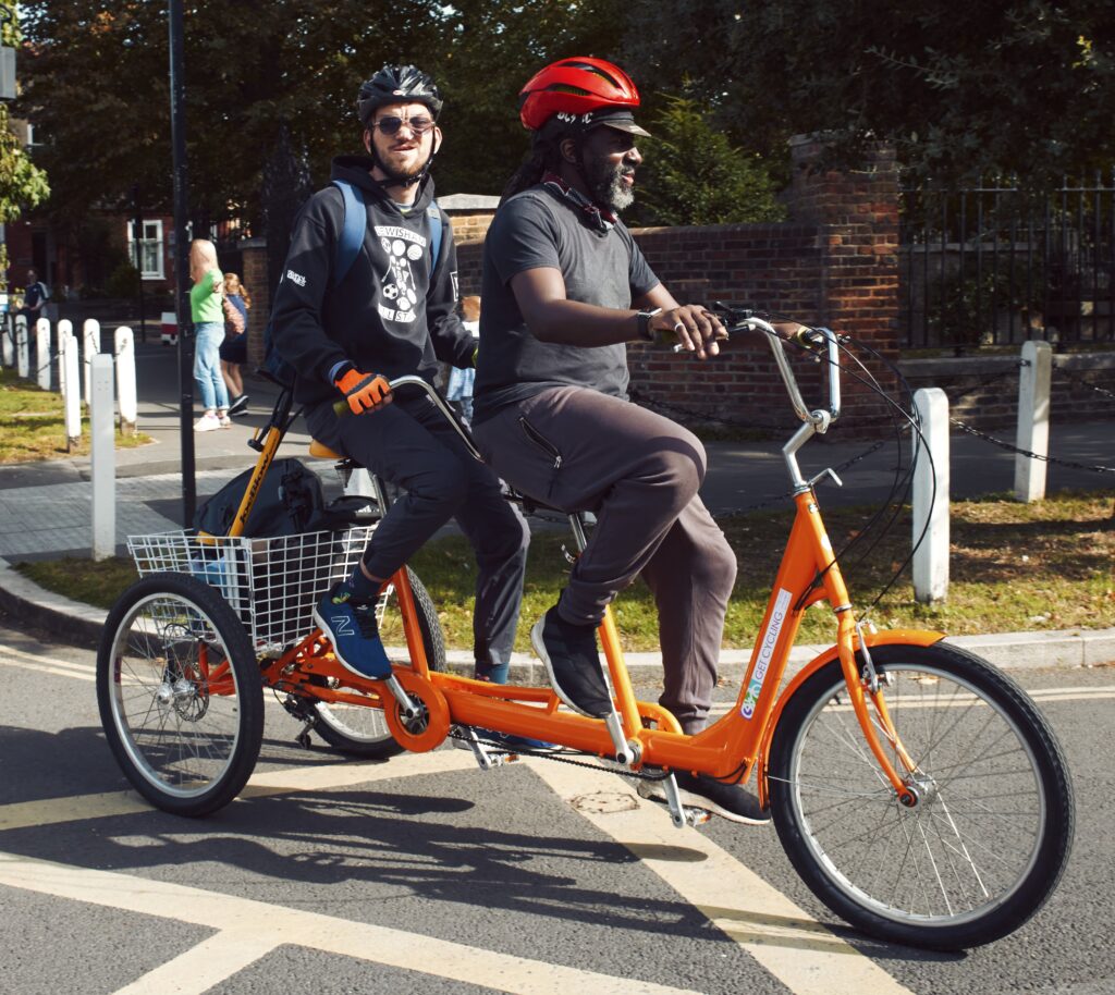 Two people riding an orange tandem tricycle through a road junction in sunshine