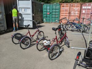 Photograph with two red upright trikes in the foreground. Behind them are some other cycles and shipping containers and an man with a high vis vest is standing with his back to the camera at the open door of a container returning the cycles for storage. 
