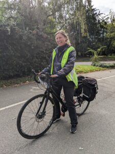 Photograph of a white woman with blonde hair in plaits standing astride a standard two-wheeled bike. She is wearing a yellow high-vis gilet over dark clothes.