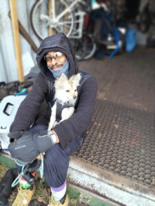 Photograph of a black man sitting on the entrance step of a cycle store. He is wearing dark clothes and has a very cute, little dog with pale fur and pointy ears sitting on his lap.