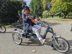Photograph of a white woman riding a recumbent trike. She is wearing dark glasses and smiling at the camera. 