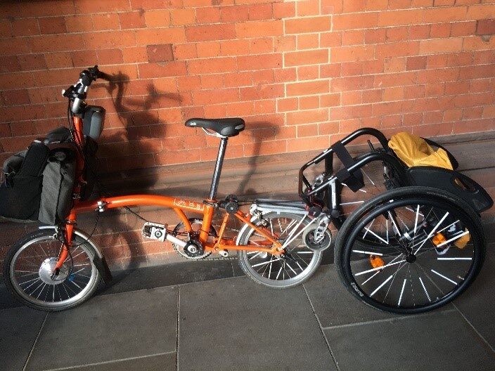 An orange Brompton with a grey front bag and e-assist has a black active wheelchair hitched to the rear pannier rack for towing