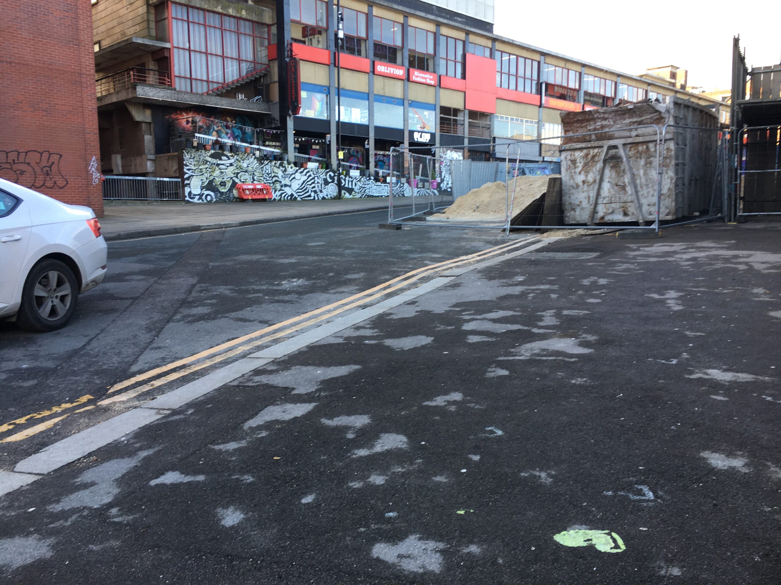 A city centre road. Photo taken from the right hand pavement looking up a significant slope. The right hand pavement is obstructed by a large skip and railings. There is a dropped kerb onto the carriageway, but no tactile paving. There is no dropped kerb to access the opposite pavement.