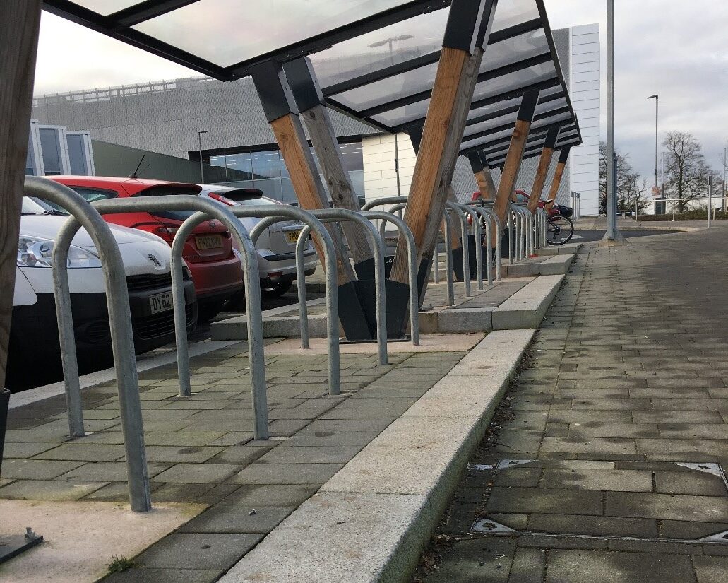 Photo of Sheffield stands under a shelter. There is a stepped kerb to access the parking, the stands are too close together and car bonnets are overhanging the cycle parking spaces.