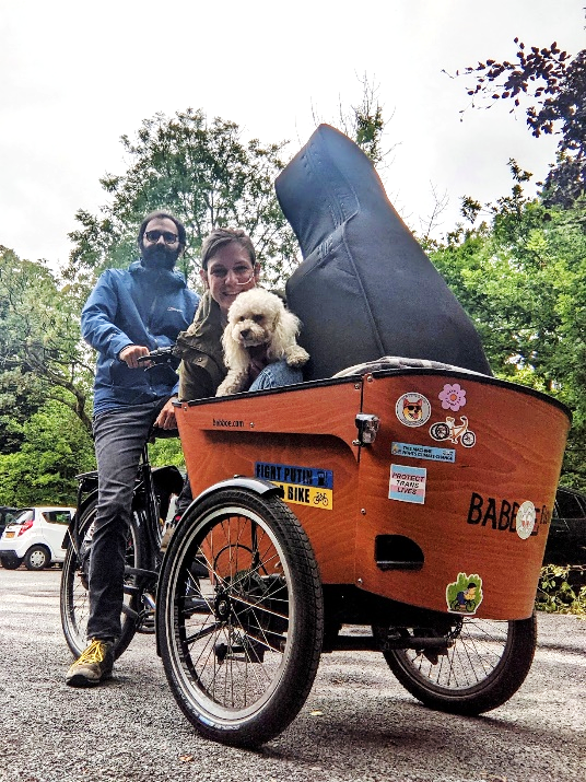 A cargo trike ridden by a white man has a white woman with a feeding tube, a poodle and a cello in the cargo box on the front