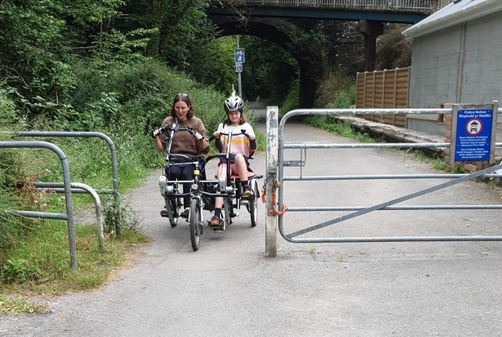A woman and child are riding a side-by-side tandem trike along a traffic-free tarmac path. They are about to pass through a gap between metal railings and a metal gate post. The gap is just wide enough for the trike.
