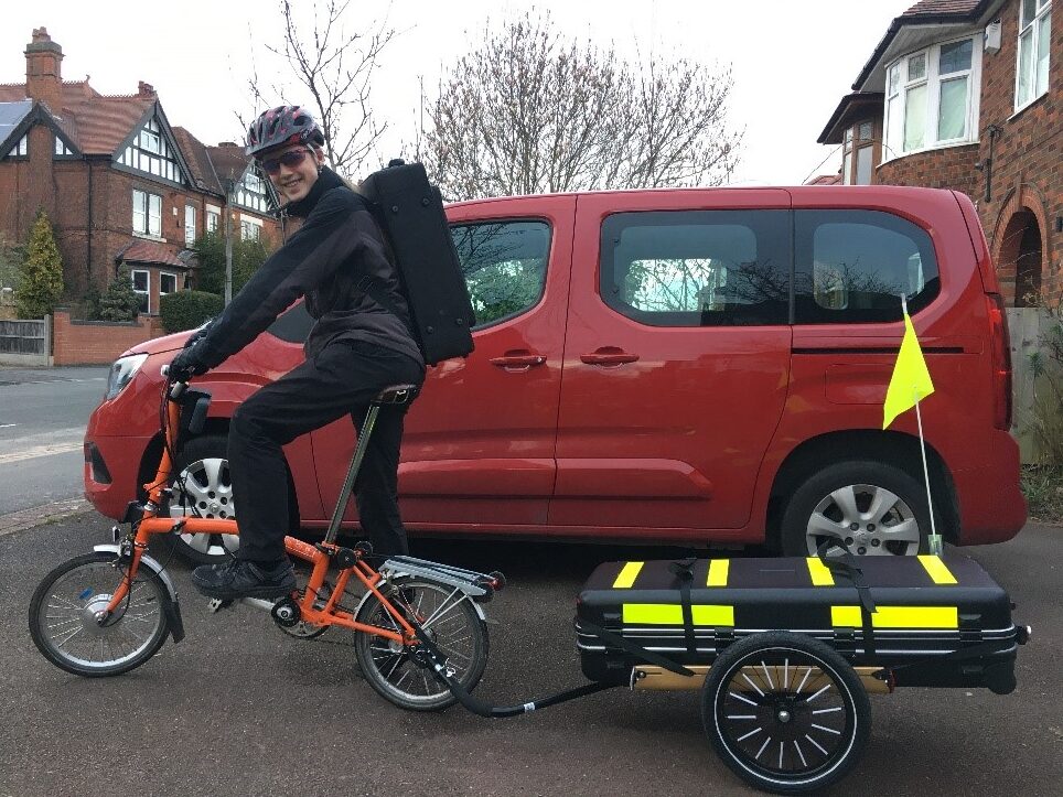 A teenager is sitting on an orange Brompton folding cycle smiling at the camera. They have an instrument case on their back and are towing a flatbed trailer with another very large instrument case strapped to it.