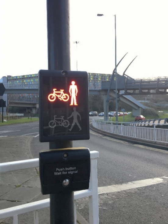 A Toucan crossing control with nearside red bicycle and man illuminated above the small metal crossing control button. There is a white railing visible behind the crossing control post.