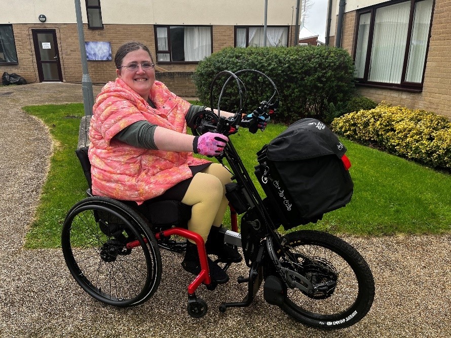 A smiling white woman wearing a pink jacket is cycling a black e-handcycle attachment on a red active wheelchair