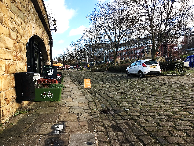 Photo of a "heritage" area with, from left to right, an entrance to a cafe obstructed by A frames and cycle parking planters; an uneven stone-slabbed pavement, no clear kerb edge onto a broad dished gutter and roadway made of large cobbles/setts, a white car parked obstructing the right hand pavement.