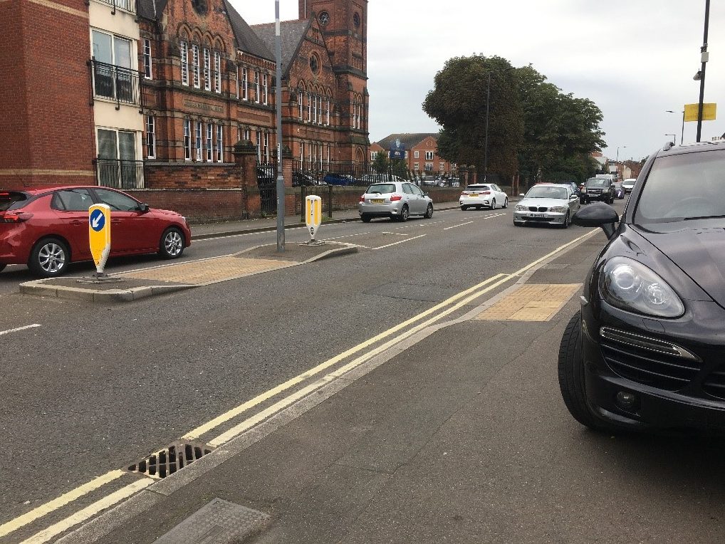 A busy urban street with 30mph speed limits, vehicles parked on the footway and a 2-stage uncontrolled crossing with buff tactile paving and a central refuge.