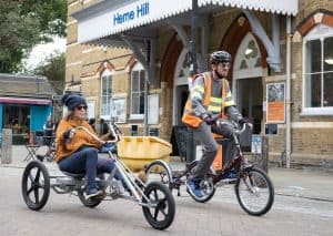 Two riders cycling on the road in front of Herne Hill train station. The man is wearing an orange hi-viz vest and black helmet, and is n a red Pashley Trike. The lady is on a grey recumbent e-trike, wearing a large comfy orange jumper and a blue bobble hat. They both look focussed and comfortable