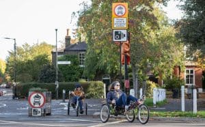 Pic of two riders cycling along abcycle lane in London. Both are on recumbent trikes. The  lady in front is in dark blue, wearing sunglasses. The lady behind has jeans, an orange jumper, and a bobblehat.