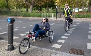 Two riders crossing the road at a cycle lane crossing point. The lady in front is on a red recumbent e-trike, wearing dark blue, and looking supremely comfortable and happy. The gentleman behind, in white helmet and hi-vis vest, is on a purple standard bicycle.