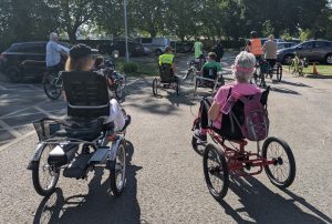 Group pic looking at the backs of 6 or 7 elderly and/or Disabled people using recumbent trikes and e-trikes. The sun is shining and they appear to be in a car park about to set off on an adventure.