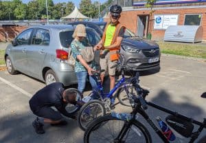 3 people are grouped around a blue tricycle. One is kneeling down to hook the chain back onto the cogs, as it has fallen off. The lady, who is the rider, is watching. The final man is gently holding her elbow and providing some balance support.