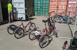 Two red tricycles are standing ready to be ridden. In the background are other tricycles and a gentleman about to enter a shipping container to see what other cycles may be available to use!