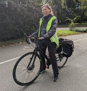 Pic of a woman standing astride her bicycle and smiling broadly- it looks like she is laughing at a joke. She has a hi-viz vest over a black waterproof jacket, black trousers, and black trainers.