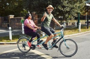 Two happy cyclists riding a light blue tandem bicycle. The male pilot has a green polo shirt, denim shorts, and a light brown pork pie hat and is giving a thumbs-up to the camera, The female stoker, in a pink t-shirt, black leggings, red trainers, and black sunglasses is smiling and basking in the sunlight.