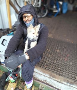 Pic of a man sitting on the edge of a storage container full of cycles. He is wrapped up warm with a dark scarf, gloves, and hooded jacket. He is smiling at the camera and in his lap is a small white dog - possibly a terrier or chihuahua mix.