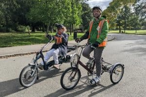 Pic of two cyclists, Em and Ben. Em is on a dark blue and grey recumbent e-trike, and is wearing jeans, a dark blue 3/4 length coat, orange/brown scarf, bobble hat and sunglasses. Ben is on an upright red Pashley trike and is wearing grey jeans, a green waterproof jacket, and an orange hi-viz vest. They are riding in a leafy green park in the sunshine.