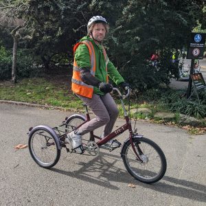 Pc of Ben riding an upright red Pashley trike in the sunshine in the park. He is wearing grey jeans, a green waterproof jacket, white trainers, and an orange hi-viz vest. 