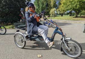 Pic of Em sitting astride her dark blue and grey recumbent e-trike in a sunny leafy park. She is wearing jeans, a dark blue 3/4 length coat, orange/brown scarf, bobble hat and sunglasses. 